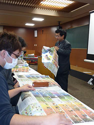 北海道新聞の見開き広告面を大学生たちに配り、模擬授業を進める山崎さん（右）
